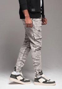 Jeans grigi sbiaditi con molteplici dettagli di cucitura, polsini elasticizzati e tasche laterali, abbinati a sneaker grigie e bianche robuste.