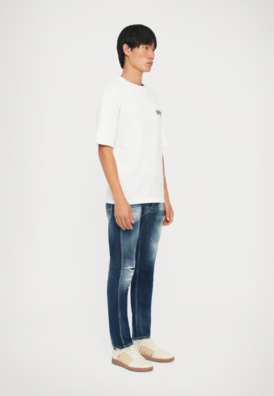 Camiseta blanca de manga corta, jeans ajustados rasgados azules, zapatillas beige con detalles marrones. Diseño simple, corte casual, sin patrones adicionales.
