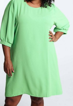 Vrouw draagt een knie-lange lichtgroene jurk met driekwart pofmouwen, één hand op de heup en de andere ontspannen langs het lichaam.