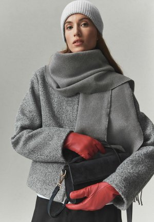 Mujer con gorro de punto gris, bufanda y chaqueta, sosteniendo un bolso negro y llevando guantes de cuero rojos sobre un fondo liso.