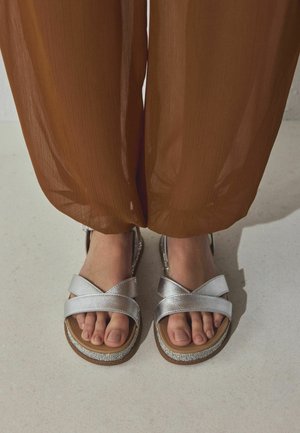 Füße in silbernen Sandalen mit gekreuzten Riemen und Strassverzierung, kombiniert mit locker sitzenden braunen transparenten Hosen, stehend auf einem hellen Boden.