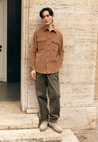 Brown corduroy shirt with two pockets, paired with olive green pants and gray boots. Standing against a textured stone wall.
