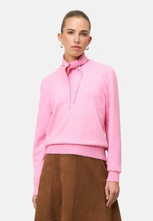 Rosa Kaschmirpullover mit geripptem Saum und einem Detail mit geknotetem Kragen, kombiniert mit einem braunen Wildlederrock, vor weißem Hintergrund.