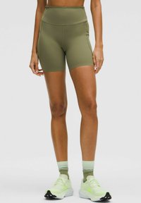 Persona che indossa pantaloni ciclistici a vita alta verde oliva, calze verdi sfumate e scarpe da corsa verde chiaro, in piedi contro uno sfondo neutro.