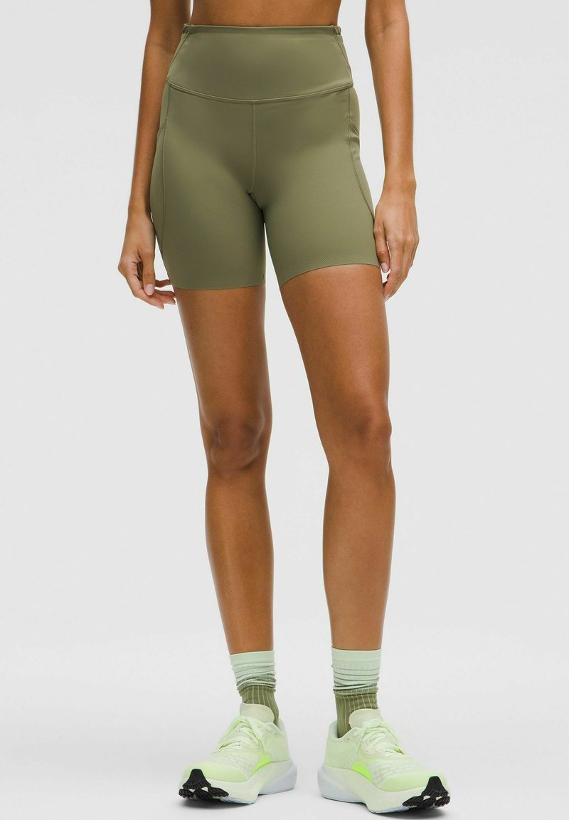 Persona che indossa pantaloni ciclistici a vita alta verde oliva, calze verdi sfumate e scarpe da corsa verde chiaro, in piedi contro uno sfondo neutro.