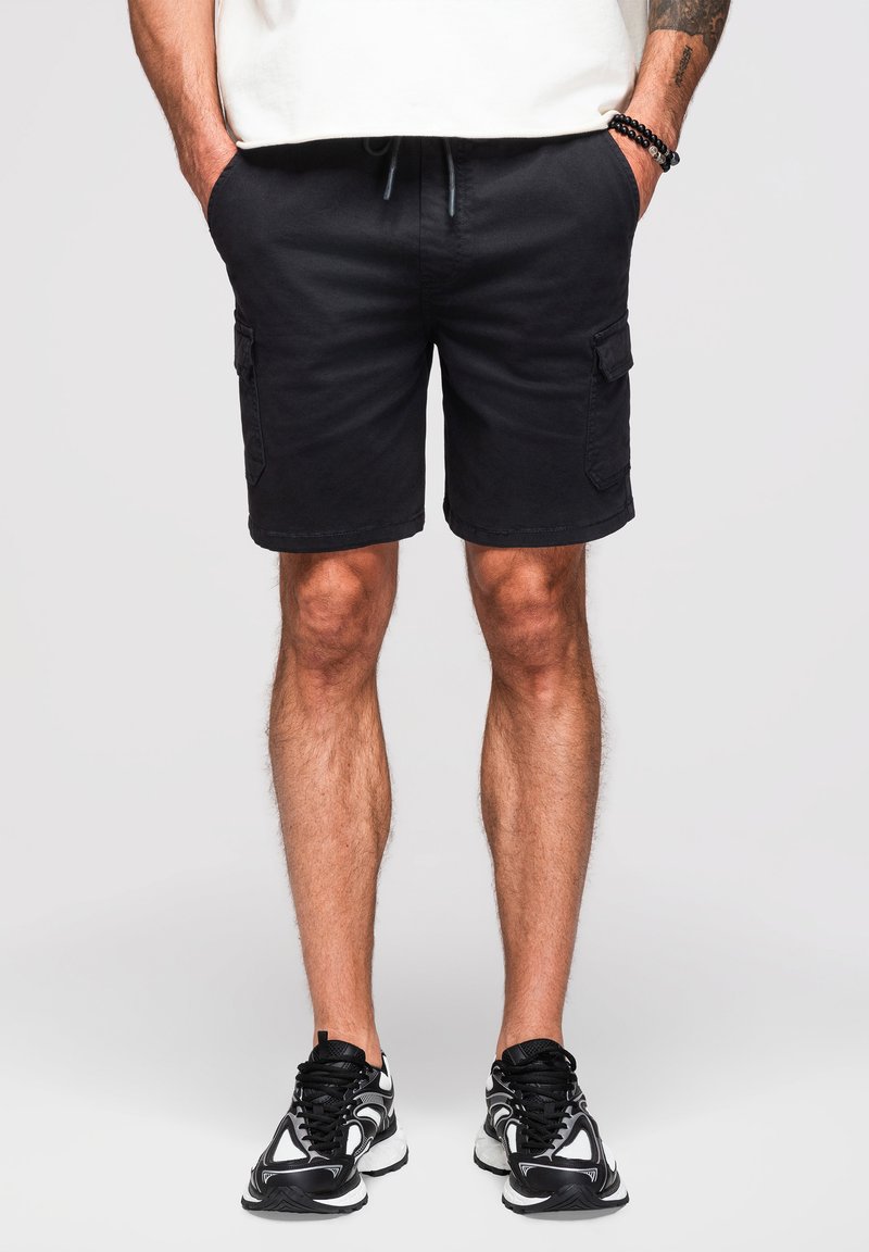 Hombre que lleva pantalones cortos cargo negros, zapatillas negras y blancas, manos en los bolsillos y una camisa blanca parcialmente visible contra un fondo liso y claro.