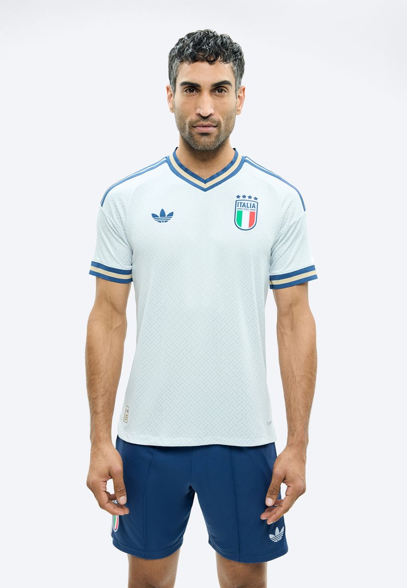 Muž v bílém a modrém italském fotbalovém dresu s logem Adidas, stojící čelem vpřed na jednobarevném světlém pozadí.