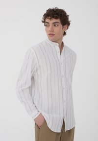 Camicia bianca a maniche lunghe con strisce verticali marrone chiaro, colletto alla mandarino e chiusura a bottoni. Tessuto leggero e traspirante, vestibilità comoda.