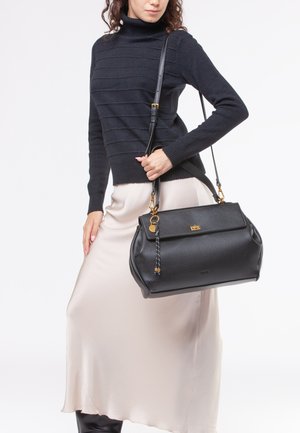 Borsa a tracolla in pelle nera con un design strutturato, hardware dorato e un accento intrecciato sulla tracolla. Indossata con una gonna in satin beige.