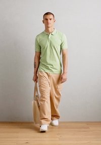 Hellgrünes Poloshirt, beiger Cargo-Hose und weiße Sneaker. Das Model trägt eine sandfarbene Tragetasche vor einem neutralen Hintergrund.