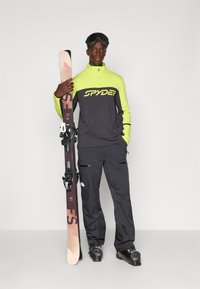 Skiklær med en grønn og grå topp med "SPYDER"-logo, svarte skibukser og et par ski, samt en snøbrille som tilbehør.