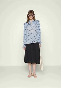 Blusa azul clara com padrão floral e colarinho com pregas, combinada com uma saia plissada preta. A modelo usa sandálias brancas e óculos de sol.