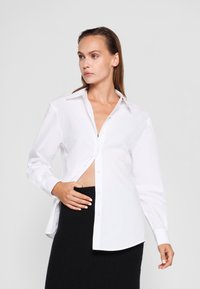 Cămașă albă cu nasturi, dintr-un material neted, având un colier clasic, mâneci lungi și o croială ușor lejeră, parțial descheiată pentru un plus de stil.