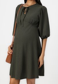 Vestido verde texturizado con mangas abullonadas, detalle de lazo en el escote tipo ojal y una silueta fluida. Combinado con un bolso cruzado marrón.