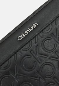 Schwarze Leder-Clutch mit einem eingravierten "CK"-Muster und einem silbernen Calvin Klein-Logo auf einer strukturierten Oberfläche. Die Kanten sind genäht.