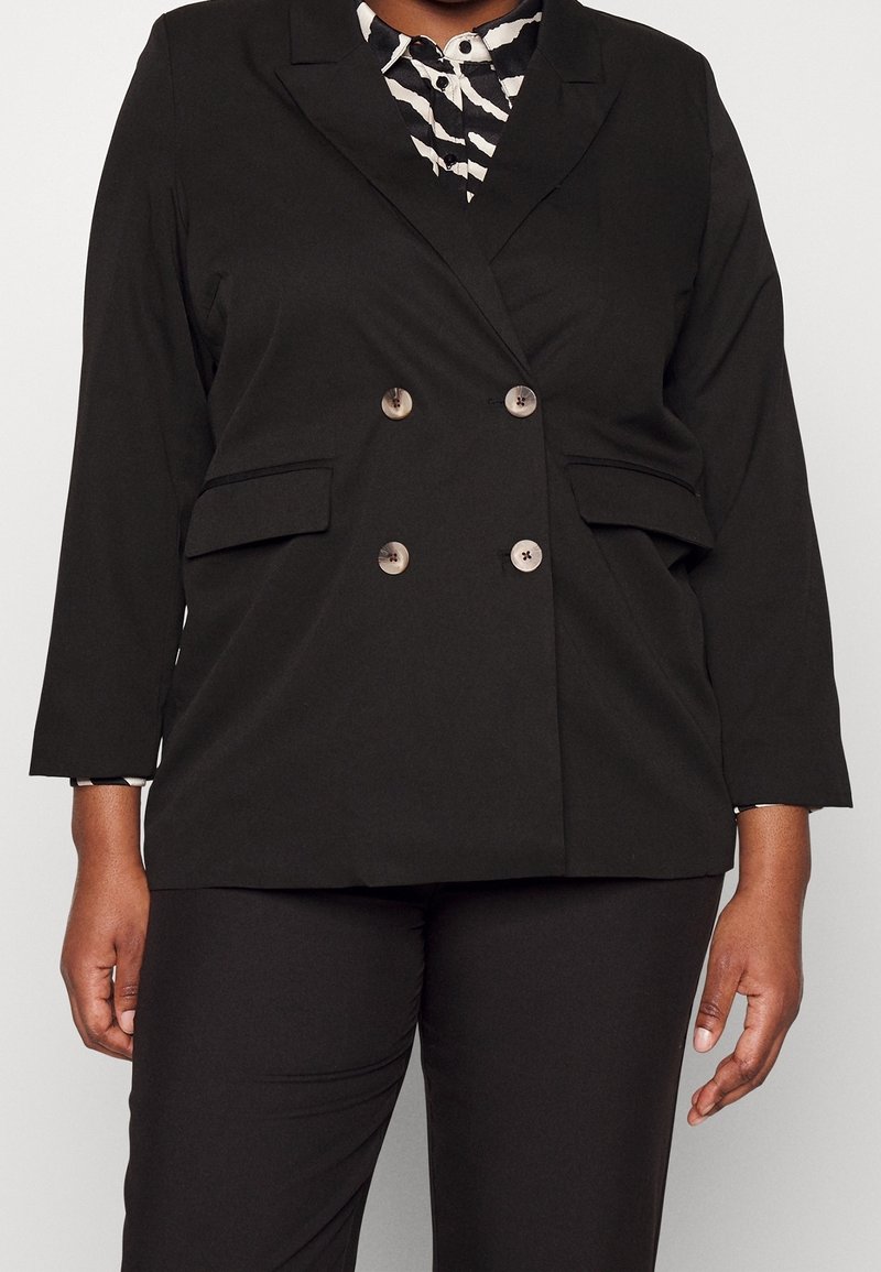 Femme portant un blazer noir croisé avec des boutons beiges, sur une chemise à motifs noir et blanc, assortie à un pantalon noir.