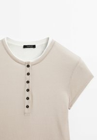 Haut à manches courtes beige à côtes avec un col rond, doté d'une bordure blanche et de six boutons noirs sur le devant. Étiquette de marque visible à l'intérieur.