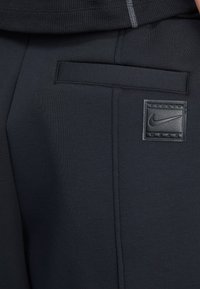 Calças de treino pretas feitas de um tecido suave, com um bolso lateral e um emblema da Nike texturizado. Ajuste justo com costuras suaves.