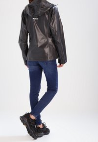 Sort vandtæt jakke med hætte, reflekterende detaljer, kombineret med mørkeblå jeans og sorte sportssko. Ren baggrund for fokus.