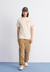 Camiseta de algodón beige con el logo "OBEY", combinada con pantalones de pana beige. El modelo lleva una gorra verde oscura y zapatillas blancas con acentos rojos.