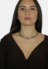 Collana choker in argento con dettaglio a cerchio, indossata sopra un top marrone a scollatura a V profonda. La collana ha una texture metallica liscia.