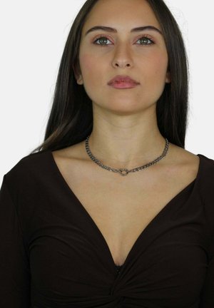 Srebrny naszyjnik choker z ogniwami w kształcie okręgów, noszony na głębokim dekolcie w brązowym kolorze. Naszyjnik ma gładką metaliczną fakturę.