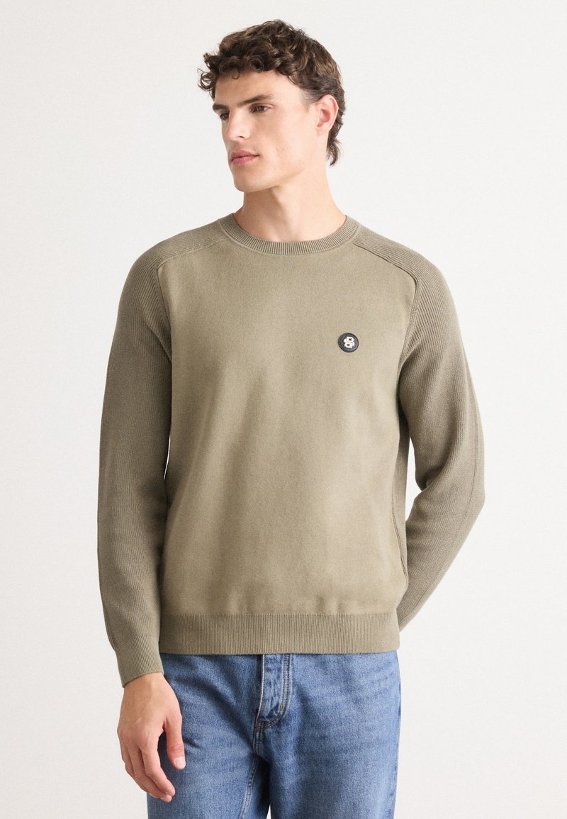 Olivgrüner Strickpullover mit rundem Halsausschnitt und geripptem Muster. Verfügt über ein schwarzes rundes Logo auf der Brust. Kombiniert mit blauen Jeans.