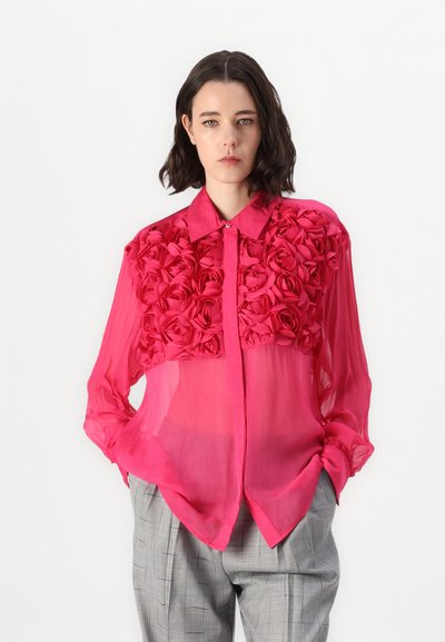 Blouse rose translucide avec un motif floral en rose texturé sur le devant, dotée d'un col classique et de manches longues, assortie à un pantalon à carreaux gris.
