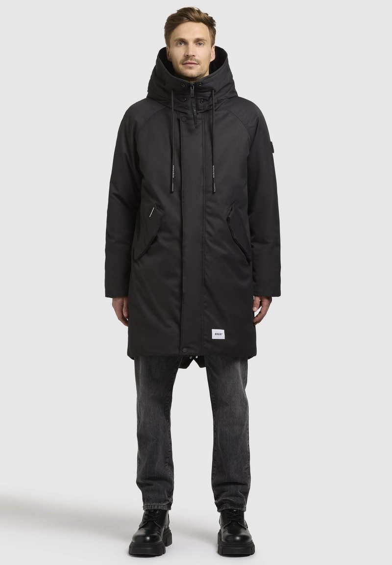 Sort parka-jakke med høj krave, snøre i hætten, to frontlommer og et logo-patch. Glat tekstur med en knælængde design.