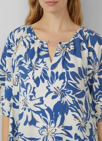 Frau trägt eine blaue und beigen Bluse mit floralem Muster, Rundhalsausschnitt und einer kleinen Knopfdetaillierung vorne.