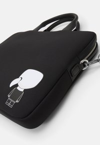 Sort neopren taske med en glat tekstur, prydet med en tegneseriefigur og en sølvfarvet lynlås. To holdbare håndtag til at bære.