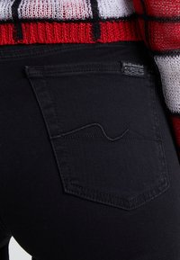 Jeans de mezclilla negra con un bolsillo trasero distintivo que presenta un detalle de costura ondulada y una pequeña etiqueta de marca. La tela tiene una textura suave.