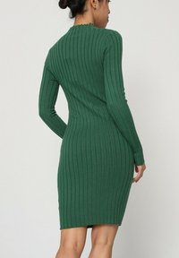 Robe ajustée et côtelée de couleur vert foncé, à manches longues et col montant, mettant en valeur un tissu texturé et une silhouette près du corps.