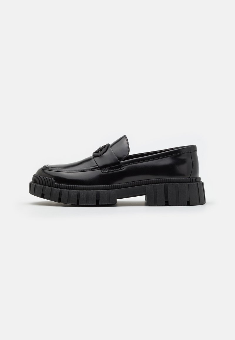 Black leather loafers with thick, ridged rubber soles. Features a rounded toe and a decorative strap with a logo accent. Smooth texture.