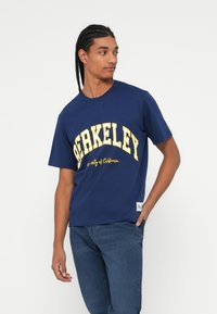 Pamučna majica tamnoplave boje s velikim žutim tekstom "BERKELEY", okruglim izrezom, kratkim rukavima i oznakom loga na donjoj strani.