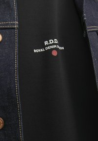 Denimu jaka ar zeltītām šuvēm, zem kuras ir melns krekls ar uzrakstu "R.D.D. ROYAL DENIM DIHON" baltā krāsā un sarkanu punktu.