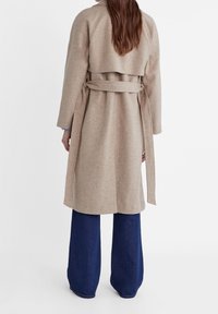 Beżowy płaszcz z wełny z paskiem w talii, bez kołnierza, z długimi rękawami. Nosi się go na niebieskie spodnie dżinsowe o szerokich nogawkach, które mają teksturowane wykończenie.