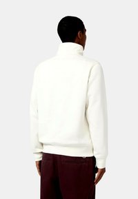 Sudadera blanca con cuello alto, corte relajado y puños acanalados. Llevada con pantalones burdeos oscuros. Textura suave, diseño minimalista.