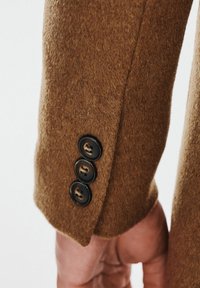 Rękaw brązowego płaszcza z włóknem wełnianym z trzema ciemnymi guzikami, miękką teksturą oraz dopasowanym krojem z czystymi detalami stębnowania.