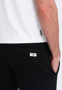 Černé joggingové kalhoty s elastickým pasem, bočními kapsami a textilní štítkem na zadní kapse. Měkký, bavlněný materiál s hladkou texturou.