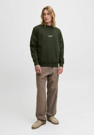 Jonge man met lang blond haar, gekleed in een donkergroene JJ Studios sweatshirts, beige broek en lichtbruine schoenen, staande voor een effen achtergrond.