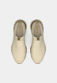 Beiget kengät, joissa on teksturoitu kangaspäällyste, pyöreä kärki ja vaalea ruskea kumipohja. Sivulla on logovempele.