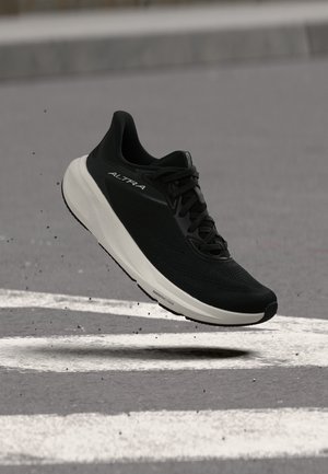 Zapatilla de running Altra en blanco y negro en el aire sobre una superficie de asfalto gris con líneas blancas pintadas.