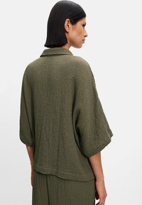 Oliwkowa, teksturowana kurtka z kołnierzem i szerokimi rękawami, charakteryzująca się luźnym krojem i delikatnie zaokrąglonym tyłem.