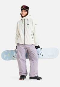 Witte waterdichte jas met zwarte rits, lichtpaarse broek, zwarte laarzen en een blauwe snowboard met een bloemenpatroon.