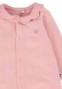 Cardigan rosa a coste con colletto a scalloped, chiusura frontale con bottoni e un piccolo emblema a forma di cuore sul lato sinistro. Tessuto morbido e texturizzato.