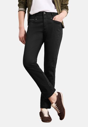 Osoba nosząca czarne spodnie o prostych nogawkach z ręką w kieszeni, w połączeniu z brązowymi zamszowymi sneakersami i oliwkowo-zieloną kurtką.