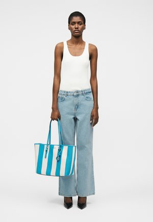 Frau steht und trägt ein weißes Tanktop sowie hellblaue, weitgeschnittene Jeans, hält eine große blaue und weiß gestreifte Tragetasche und trägt schwarze High Heels.