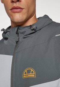 Hall värvi kapuutsiga jakk, millel on helehall paneel. Omab reguleeritavat paelakinnitust, lukku ning oranži Ellesse logo rinnal.