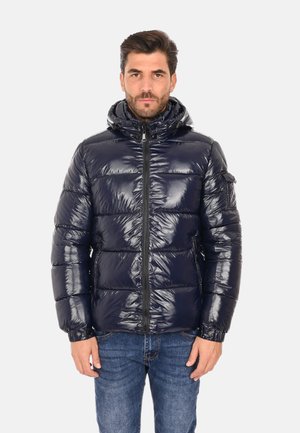 Giacca a vento blu navy con texture trapuntata lucida, cappuccio attaccato, zip frontale e polsini elastici. Indossata sopra jeans di denim.